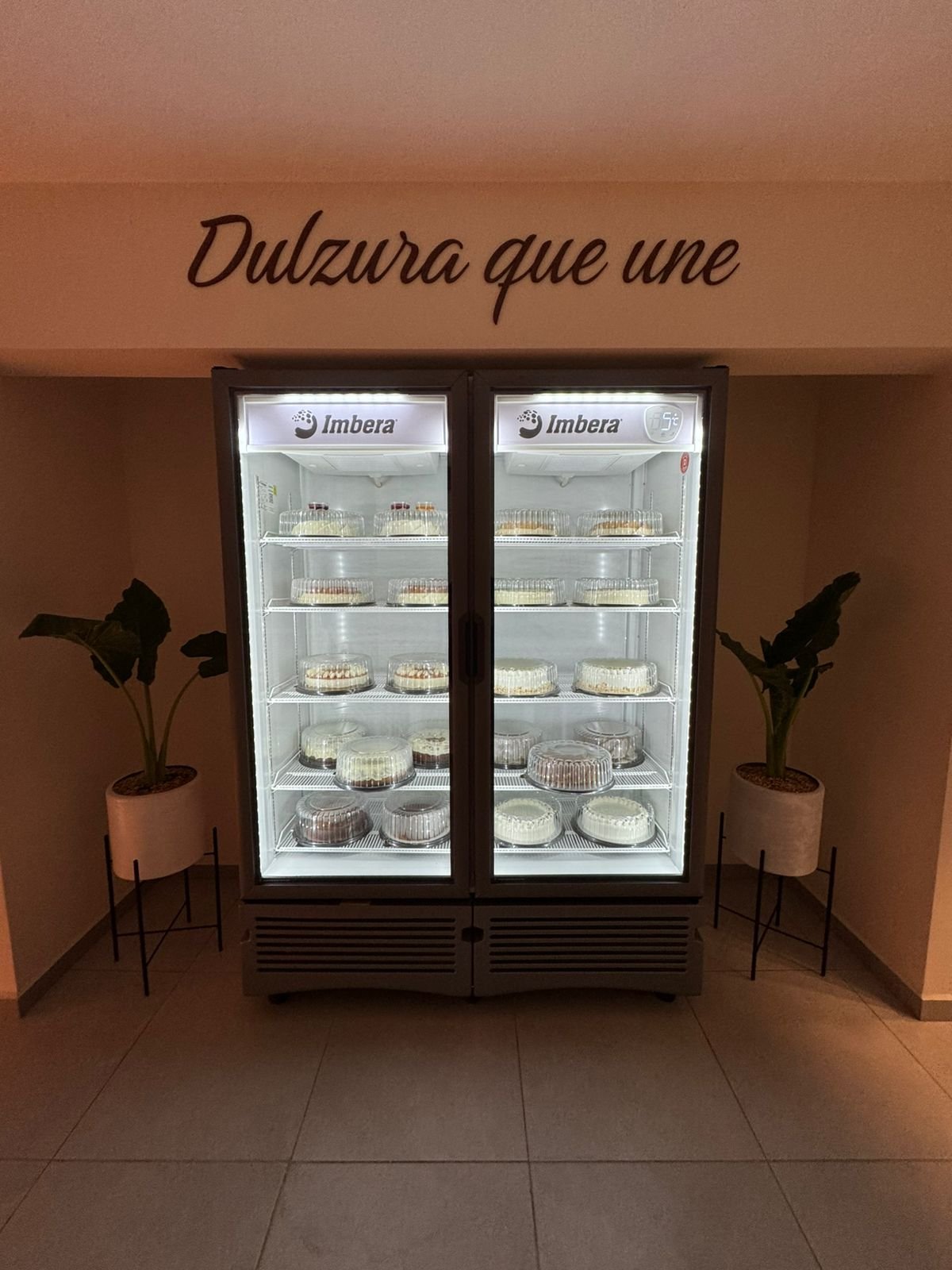Two Imbera refrigerators displaying baked goods in a pastry shop with plants on each side and Spanish text on the wall