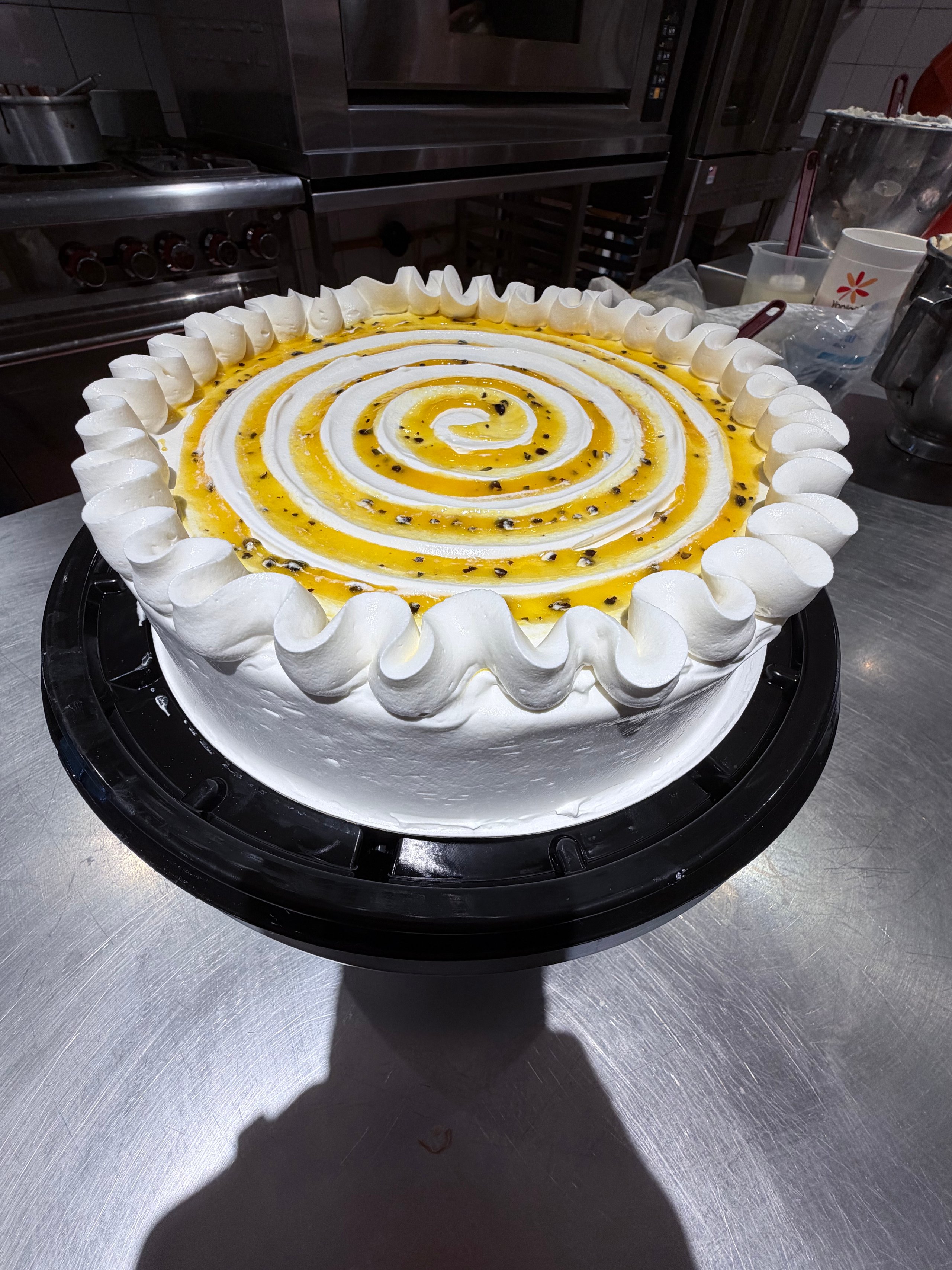 A round cake with white frosting piped border, yellow and white spiral topping, displayed on a black cake base in a kitchen